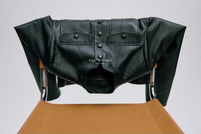 Black leather jacket by Rag & Bone displayed on a brown leather and chrome chair against a neutral studio backdrop.