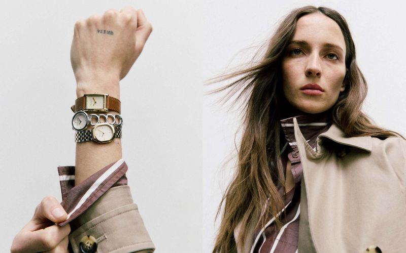 Woman poses with vintage watches accessorizing a tan trench coat and striped collared shirt in a studio shot, against a plain white background.