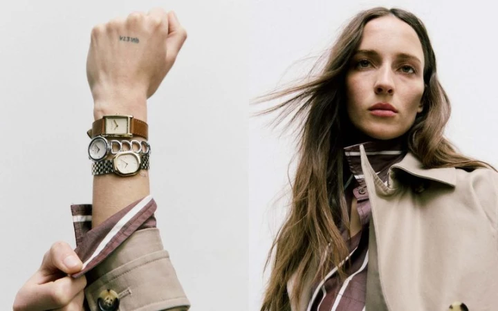 Woman poses with vintage watches accessorizing a tan trench coat and striped collared shirt in a studio shot, against a plain white background.