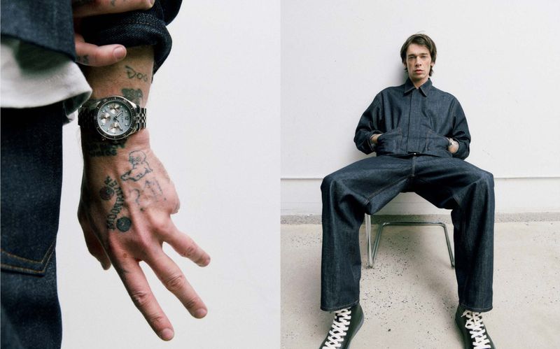 Model wears a full denim outfit while sitting on a stool near a tattooed hand wearing a watch, set against a plain white background.