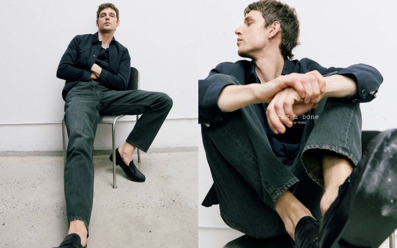 A casually dressed adult male sits on a chair in a studio, wearing jeans, a dark top, and loafers. He exudes a relaxed, cool vibe.