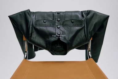 Black leather jacket draped over a brown leather chair against a neutral grey studio background