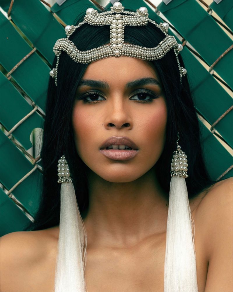 Close-up of a woman with dark hair and light skin wearing an ornate beaded headpiece and earrings, against a woven green backdrop.