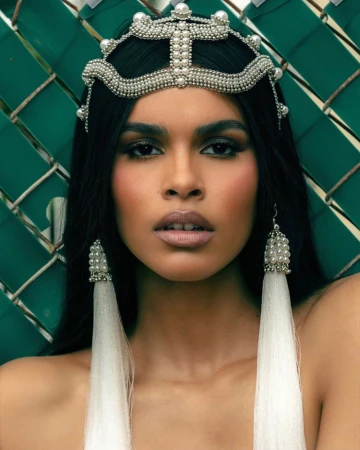 Close-up of a woman with dark hair and light skin wearing an ornate beaded headpiece and earrings, against a woven green backdrop.