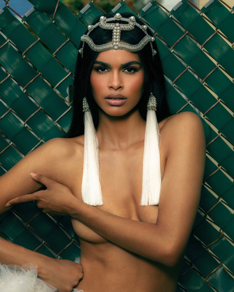 Striking portrait of a woman with a jeweled headpiece, statement earrings, and partially covered torso against a green chain-link background.