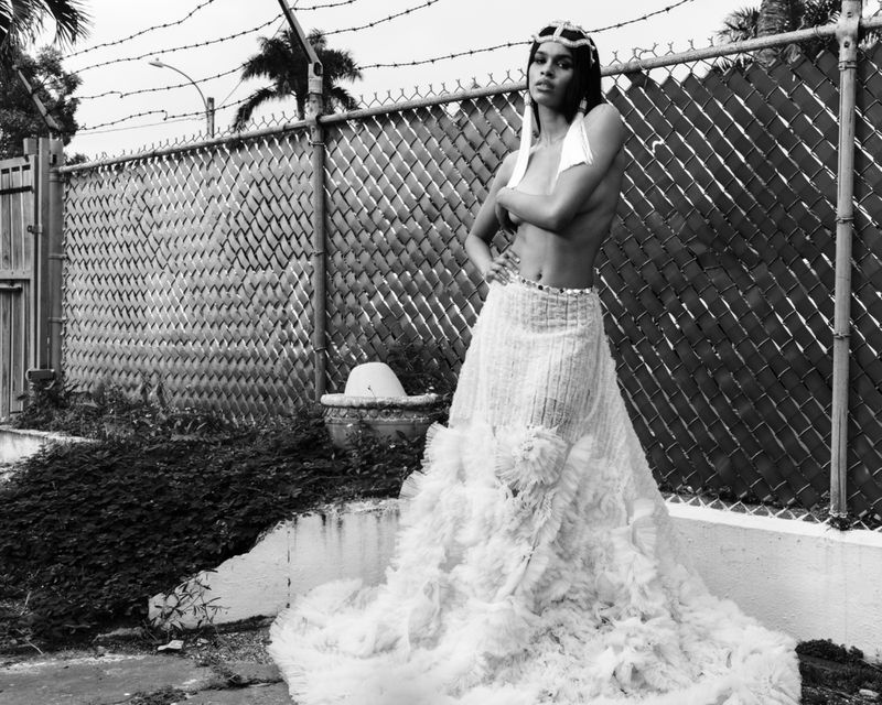 Monochrome portrait of a woman in a ruffled skirt and jeweled headpiece posing near a chain-link fence with palm trees in the background.