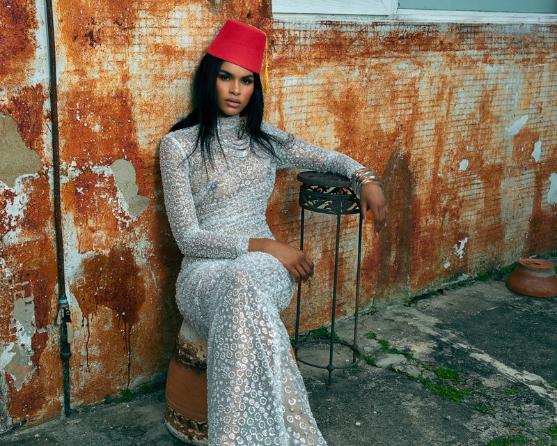 Elegant woman in a white lace gown and a red fez sits outdoors against an aging wall, exuding a refined and mysterious aura.