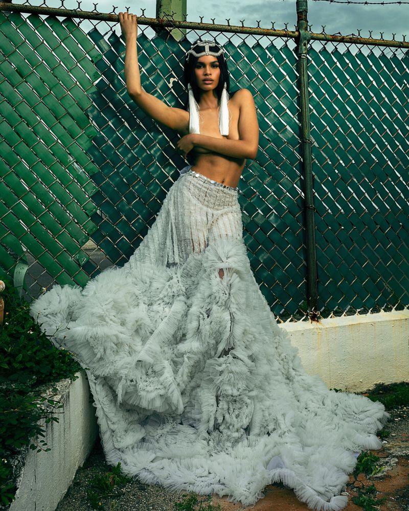 A woman poses in a flowing ruffled skirt against a chain-link fence, accented by a jeweled headband and long, white earrings in an outdoor setting.