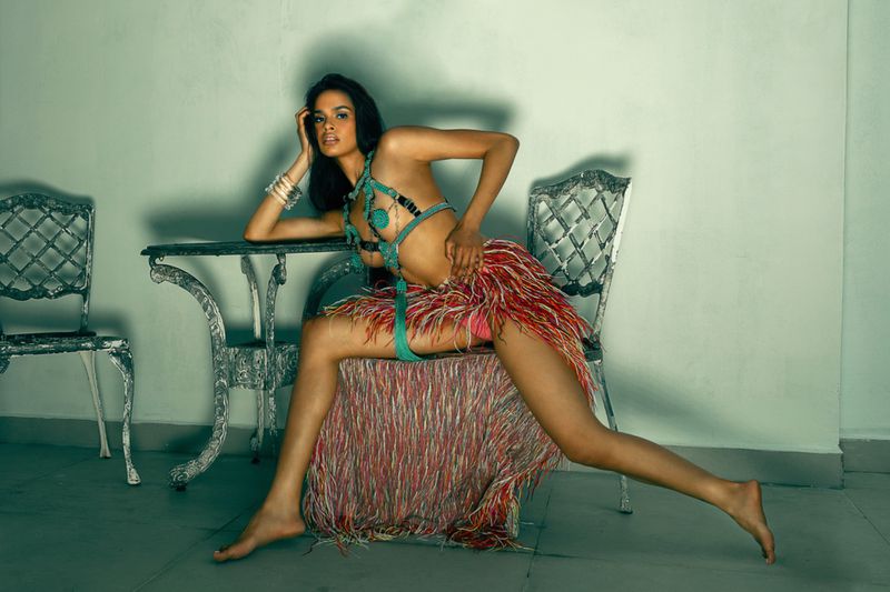A woman poses in a studio wearing a turquoise bikini and feathered skirt, seated on a chair behind a table, gazing at the camera.