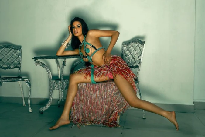A woman poses in a studio wearing a turquoise bikini and feathered skirt, seated on a chair behind a table, gazing at the camera.