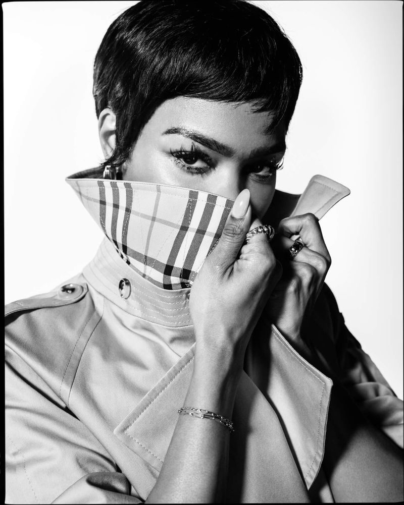 Black and white portrait of a woman with short hair, wearing a trench coat with a plaid collar, covering part of her face with the collar.