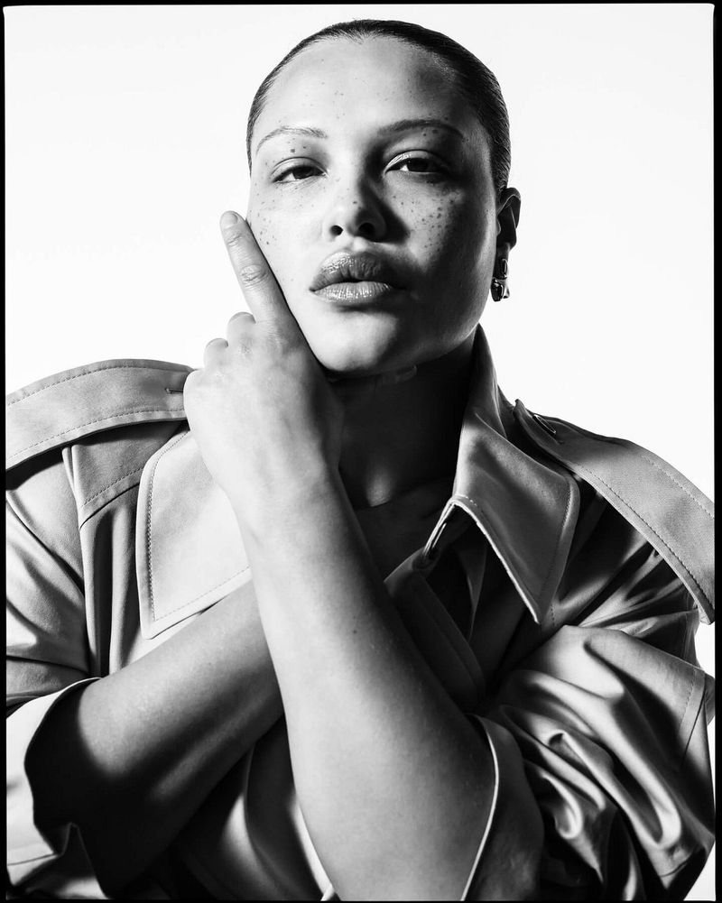 Monochrome portrait of a woman with short hair wearing a trench coat, her hand near her face, against a bright, white backdrop.