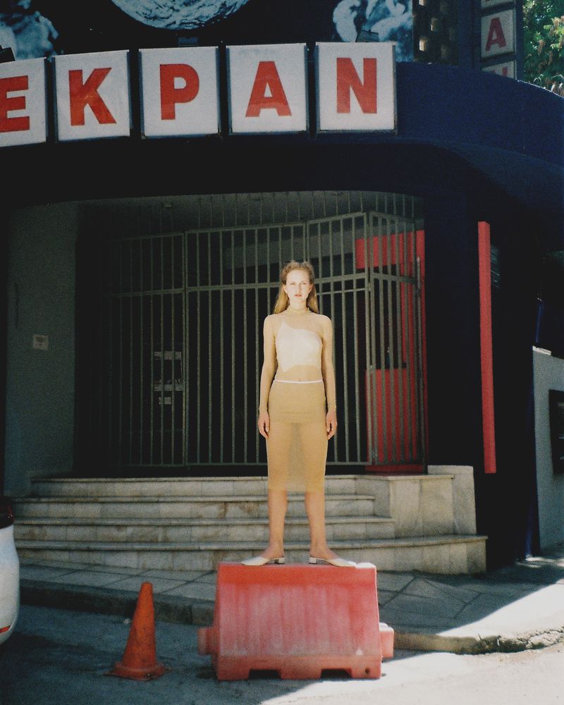 Young woman in sheer beige outfit standing on a red traffic barrier in front of a building with stairs and red signage.