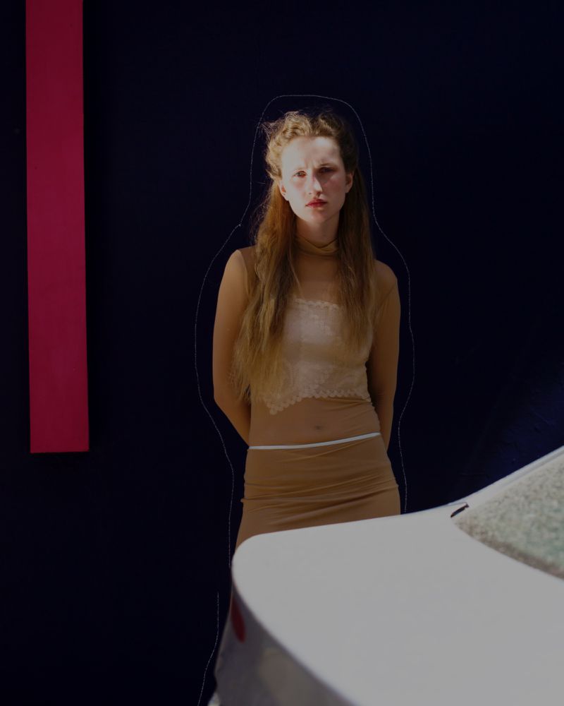 Blonde woman in neutral-toned outfit poses next to a modern white car against a dark backdrop with a vertical pink stripe.