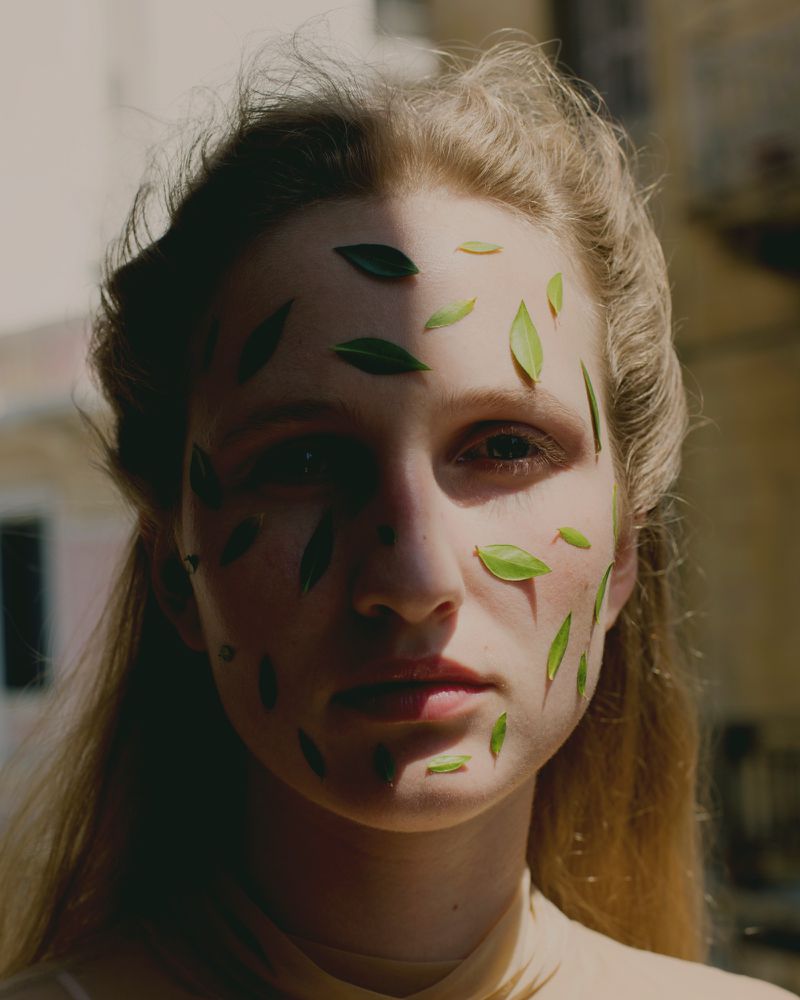 Portrait of a blonde woman with green leaves arranged on her face, partially shadowed with natural outdoor lighting.