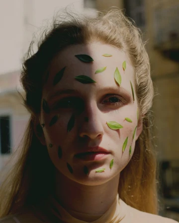 Portrait of a blonde woman with green leaves arranged on her face, partially shadowed with natural outdoor lighting.