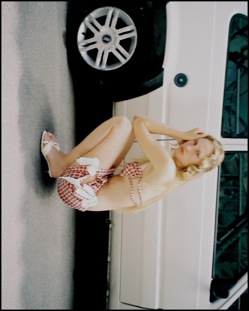 Blonde woman poses next to a white van wearing a gingham bikini and high heels with her knees to her chest on asphalt outdoors.