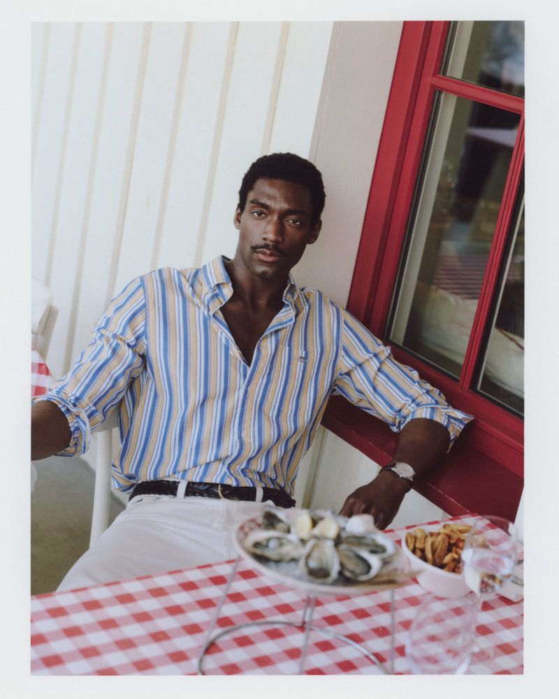 Handsome African-American man poses at an outdoor restaurant table with oysters and fries, wearing a striped shirt and white pants.