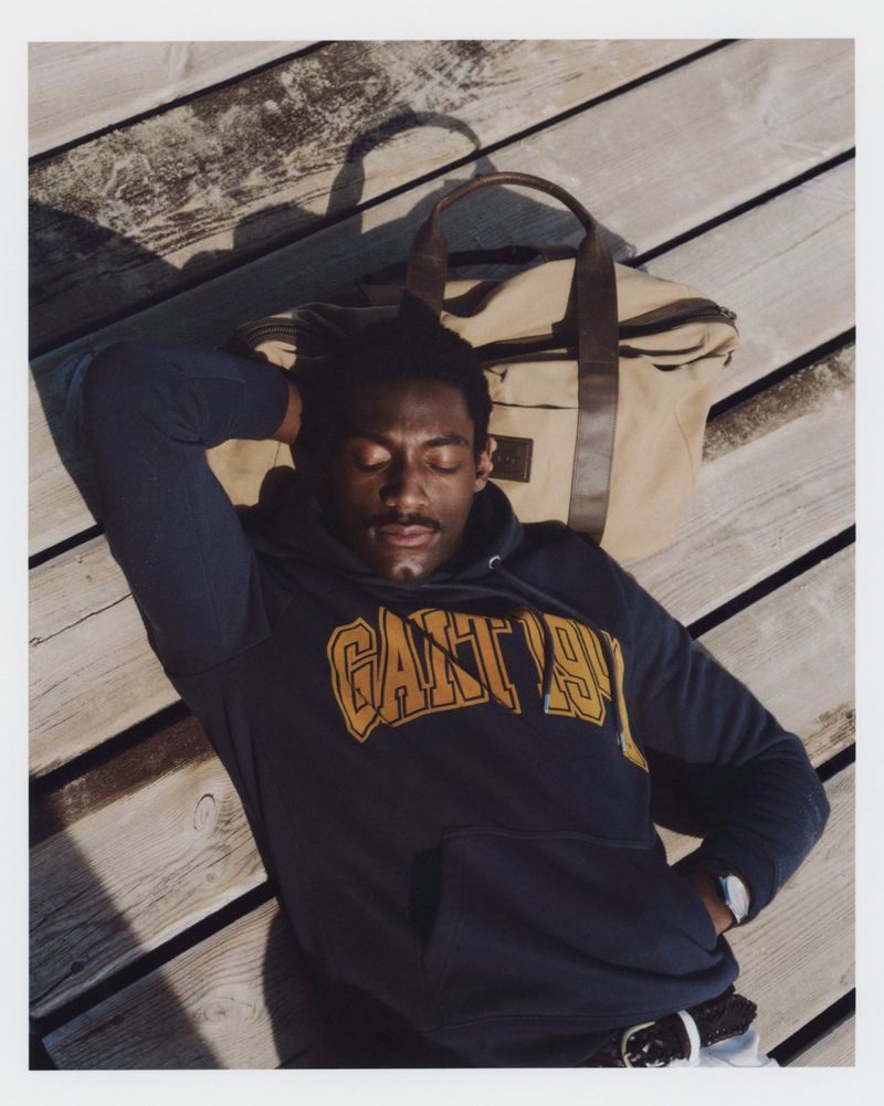 Man in Gant hoodie relaxes on wooden dock with duffel bag, eyes closed in sunlight, wrist watch, and navy, brown, and tan colors.