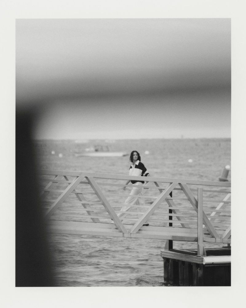Monochrome shot of a woman in a black and white shirt walking on a pier; a boat is visible in the distance over the water.