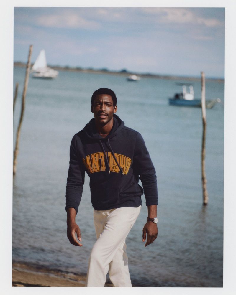Outdoor portrait of a Black male model wearing a navy hoodie and cream-colored pants with boats in the background by the water.