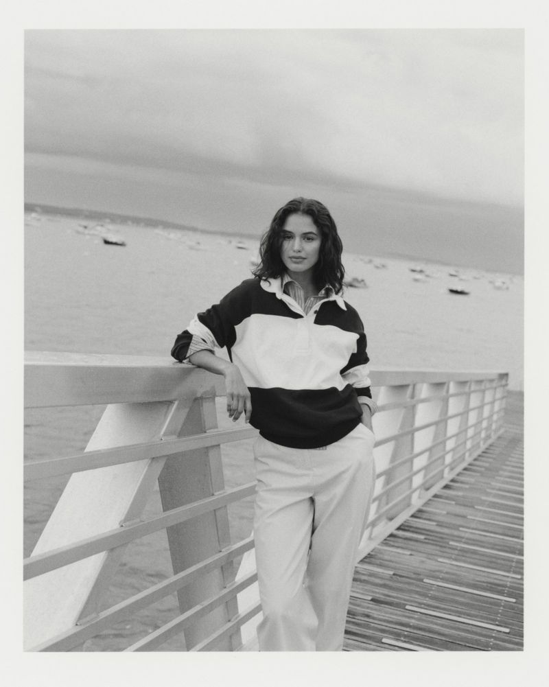 Black and white portrait of a woman with dark curly hair in a color-blocked shirt, standing near a wooden railing by a body of water.