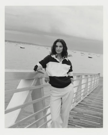 Black and white portrait of a woman with dark curly hair in a color-blocked shirt, standing near a wooden railing by a body of water.