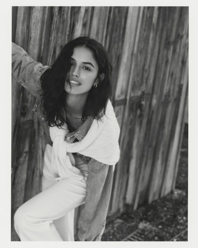 Portrait of a young woman with dark hair in denim jacket and white pants, standing outdoors in front of a wooden wall and gravel ground.