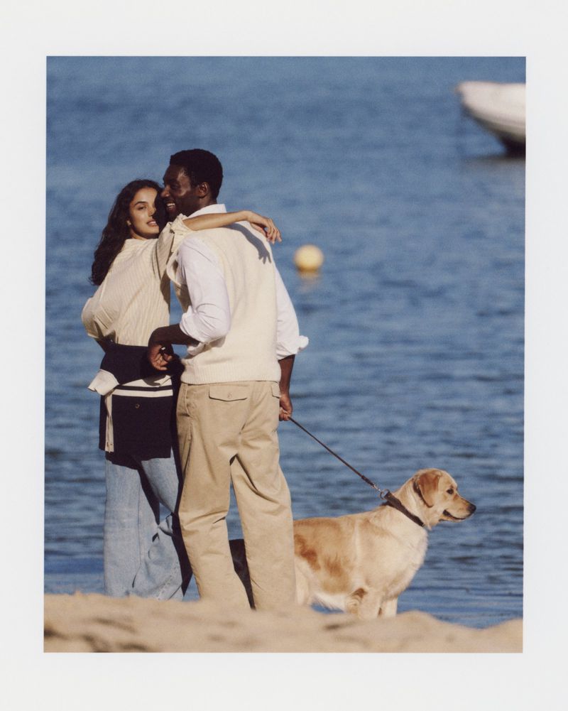 Couple with a golden retriever standing on a sandy beach near the blue ocean water, with a boat visible in the background.