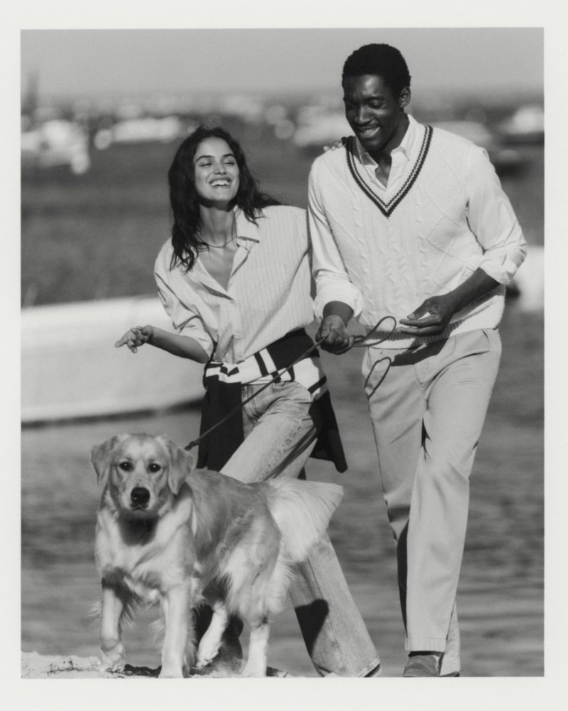 An adult man and woman walk a golden retriever dog near a harbor on a bright day, captured in a black-and-white outdoor shot.