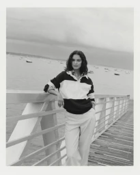 Model leaning on a wooden boardwalk railing against an overcast waterfront background wearing a striped rugby shirt.