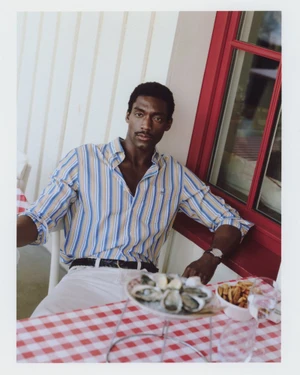 Model seated outdoors at a checkered table wearing a blue and yellow striped shirt for Gant