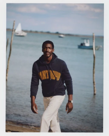 A Black man walking along a shoreline wearing a navy hoodie and light trousers for Gant Spring Summer 26.