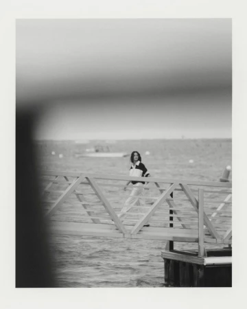 Model walking on a pier over water wearing a collared top and light trousers, captured in monochrome style.