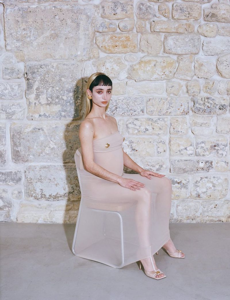 Young woman with bangs and light skin sits in a sheer dress on a transparent chair in front of a textured stone wall indoors.