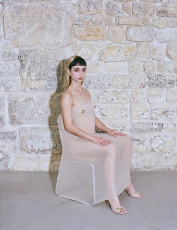 Young woman with bangs and light skin sits in a sheer dress on a transparent chair in front of a textured stone wall indoors.