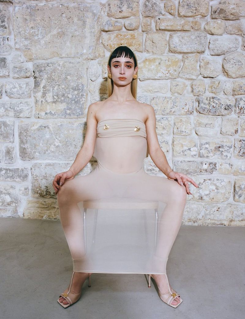 A slender woman with bangs poses in a beige tube dress and high heels, seated on a transparent chair against a stone wall backdrop.