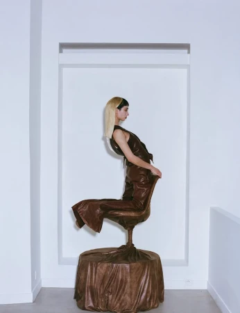 A woman in a sleeveless brown leather suit sits in a leather chair, covered with draped leather fabric, in a minimal indoor setting.