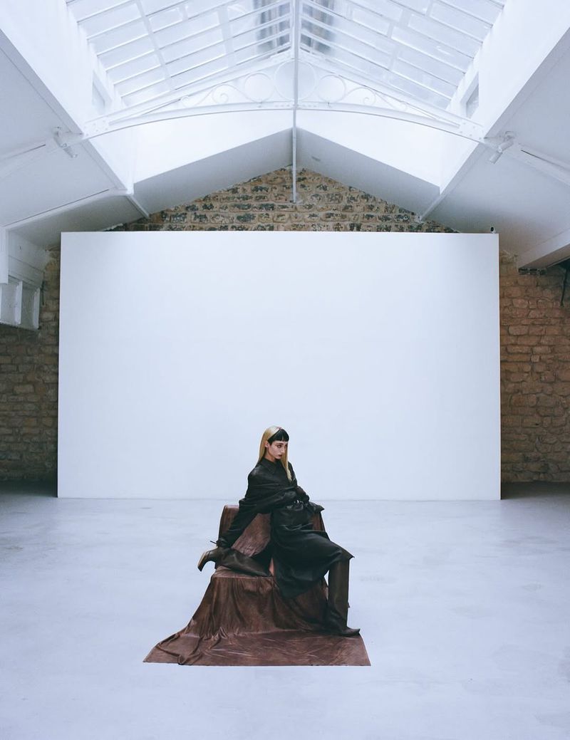 Adult female fashion model in dark clothing posing in an indoor photo studio with neutral color palette and ample natural overhead lighting.