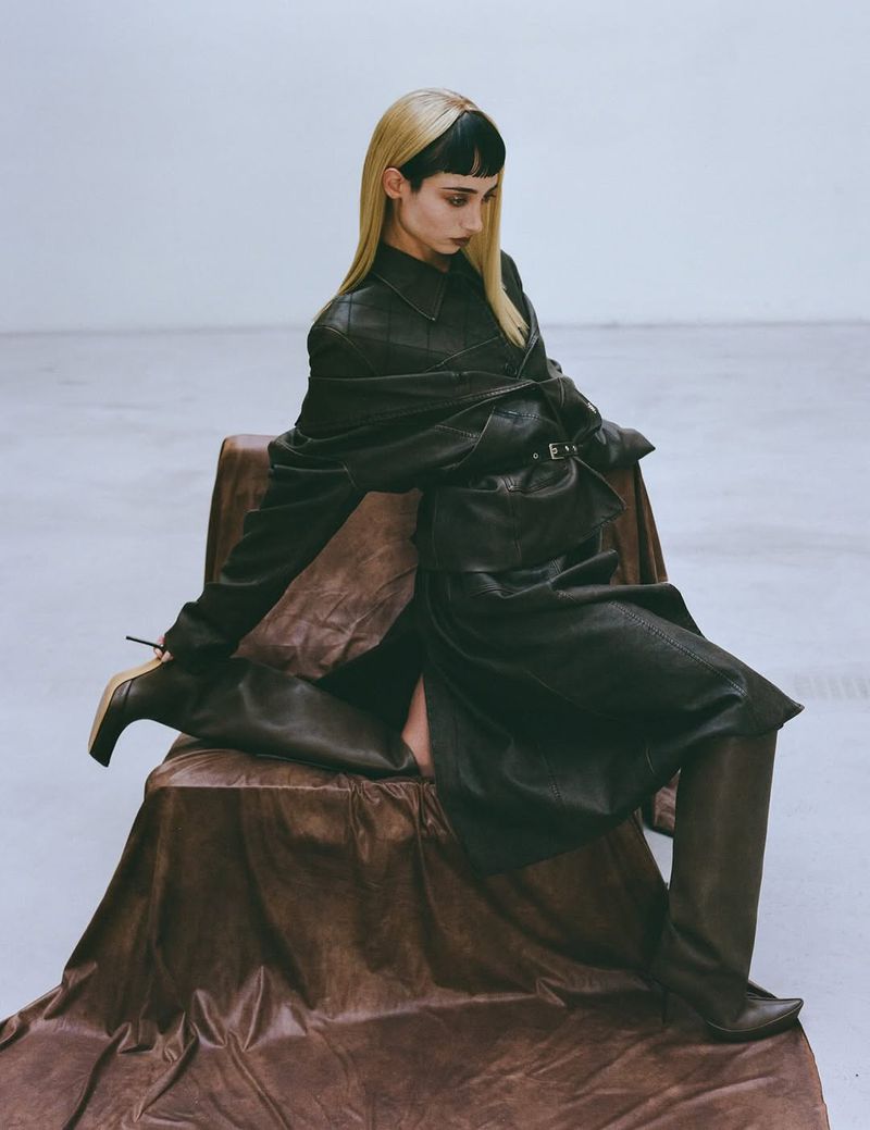 A solitary woman with blonde hair and bangs sits gracefully on a chair draped with brown fabric, wearing a dark jacket and boots in a stark studio.