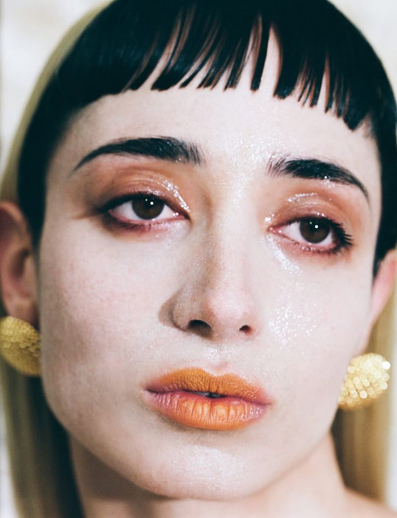 Close-up portrait of a woman with black and blonde hair, wearing gold earrings and bronze makeup, with a focus on beauty and fashion.