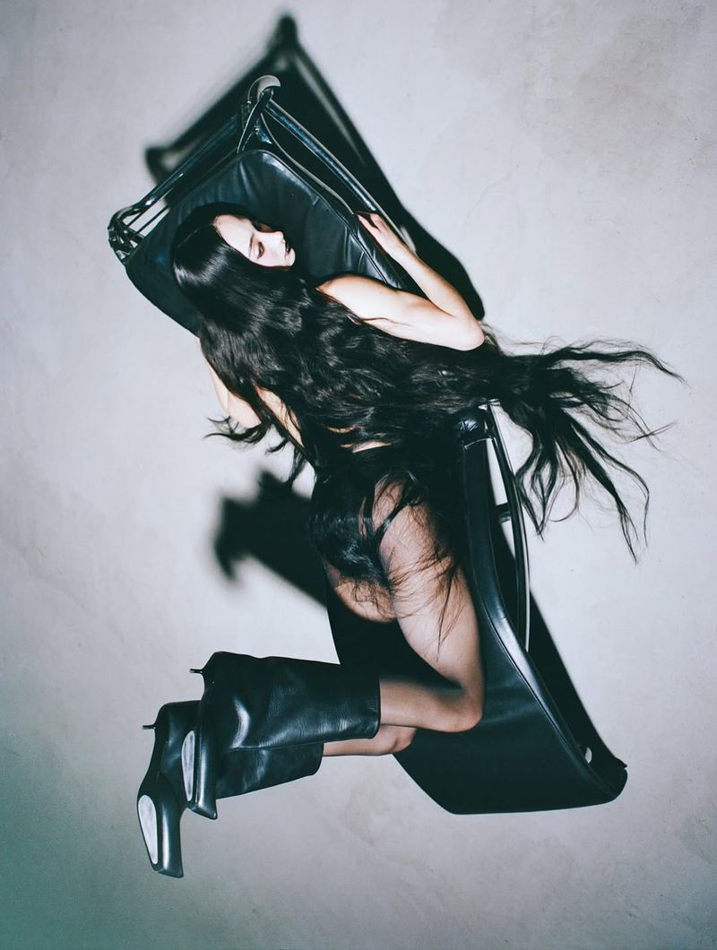 Woman with long dark hair poses in black boots and clothing, reclining on a black chair against a pale studio backdrop in a fashion shot.