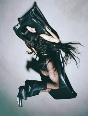 Woman with long dark hair poses in black boots and clothing, reclining on a black chair against a pale studio backdrop in a fashion shot.