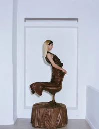 Model in brown leather outfit seated strangely on a matching covered chair against white walls