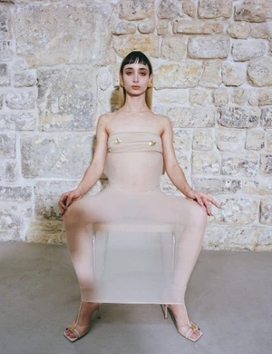 Woman in sheer beige jumpsuit seated on a transparent chair against an exposed stone wall.