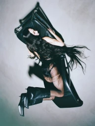 Woman with long dark hair lying on a black leather lounge chair wearing black boots against a textured grey background.