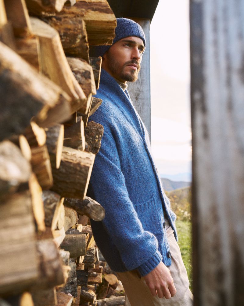 Man in blue cardigan and beanie leans against a stack of firewood. Natural light illuminates the outdoor scene, emphasizing texture.