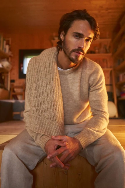 Indoor portrait of a Caucasian adult male with dark hair and a beard, wearing a sweater and scarf, sitting on a wooden surface.