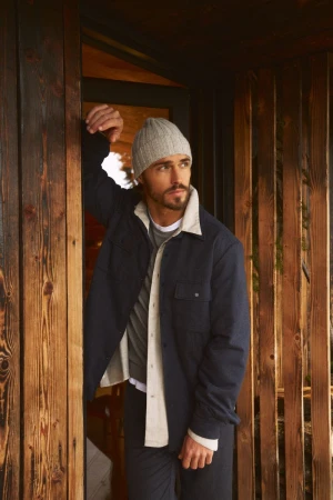 A handsome adult male with a beard and beanie stands in a doorway with wooden planks, looking out into the distance in a casual pose.