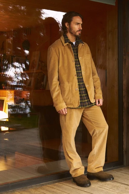 Man in a tan corduroy suit and plaid shirt leans against a glass wall in a warm indoor setting with diffused natural light.